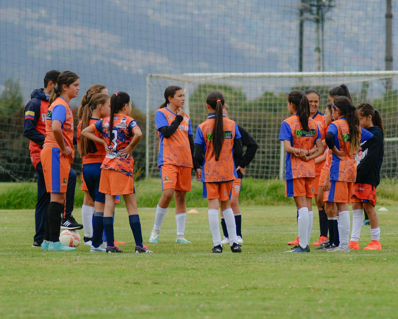 A group of young girls in soccer sportswear huddle during practice on an outdoor field.